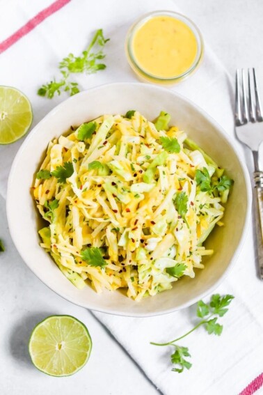 Jicama slaw in a bowl with lime halves, cilantro, dressing and a fork on the side.