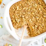 Dish of baked zucchini bread oatmeal topped with chopped walnuts. A wooden spoon is in the dish for serving. Sliced zucchini and walnuts are on the table around the dish.