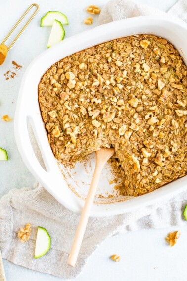 Dish of baked zucchini bread oatmeal topped with chopped walnuts. A wooden spoon is in the dish for serving. Sliced zucchini and walnuts are on the table around the dish.