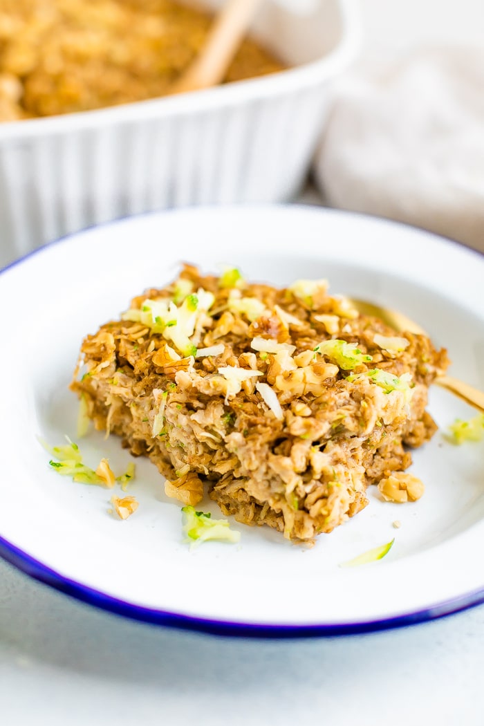 A slice of zucchini bread baked oatmeal topped with some grated zucchini and chopped walnuts on a blue and white plate with a gold spoon.