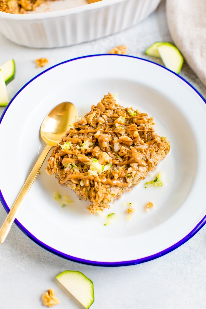A slice of zucchini bread baked oatmeal topped with a drizzle of almond butter and chopped walnuts on a blue and white plate with a gold spoon.