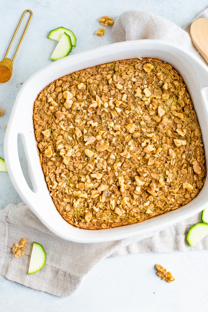 Dish of baked zucchini bread oatmeal topped with chopped walnuts. Sliced zucchini and walnuts are on the table around the dish.