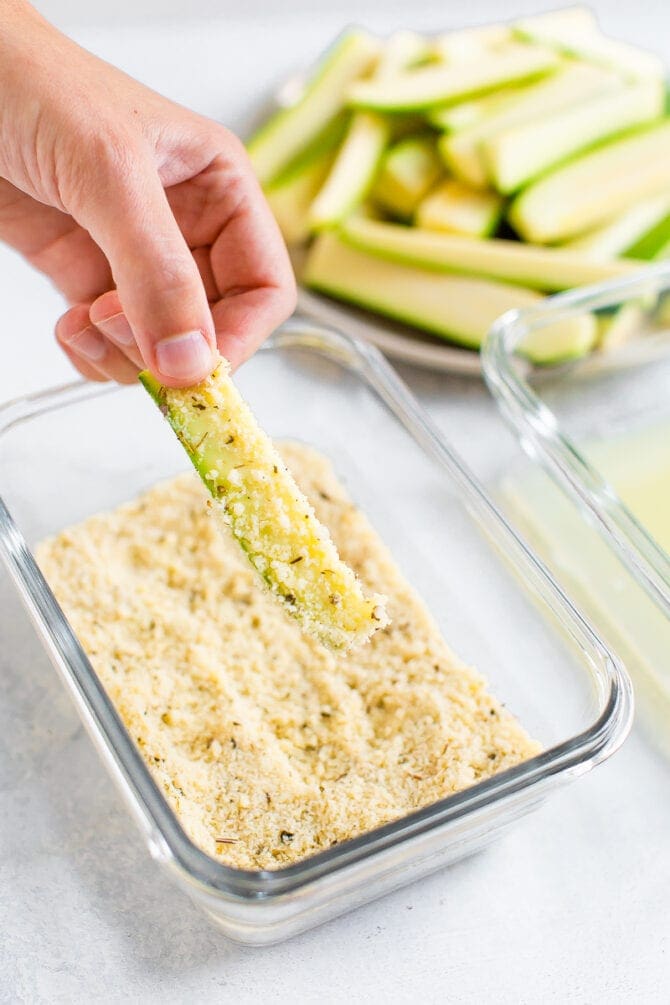 Hand holding a piece of zucchini and coating it in an almond flour and parmesan coating.