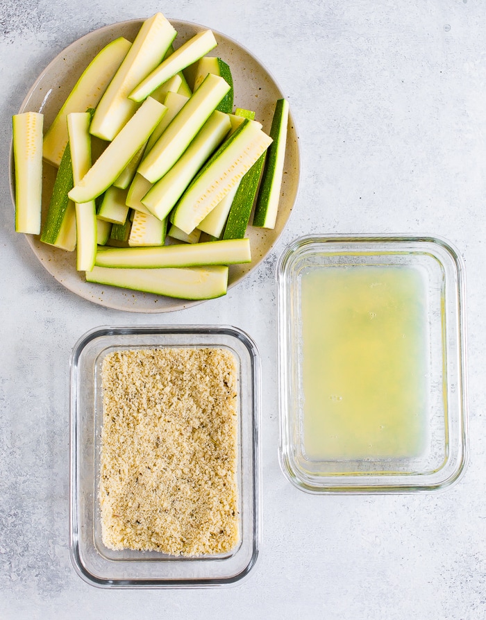 Staged ingredients for making baked zucchini fries: a plate of chopped zucchini, a dish with almond flour and parmesan coating, and a container with egg whites.