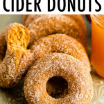 Apple cider donuts on a tray. One had a bit taken out of it, and a mason jar of cider is beside the donuts.