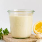 Jar of tahini sauce. Lemon, parsley and garlic cloves are around the jar.