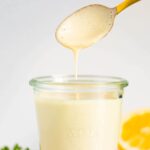 Jar of tahini sauce with a spoon drizzling some from the jar. Lemon, parsley and garlic cloves are around the jar.