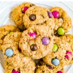 Plate of gluten-free peanut butter monster cookies with color candies in them. A dish towel and glass of milk are on the side.