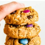 Hand holding a stack of three gluten-free peanut butter monster cookies made with oats and colored candies.