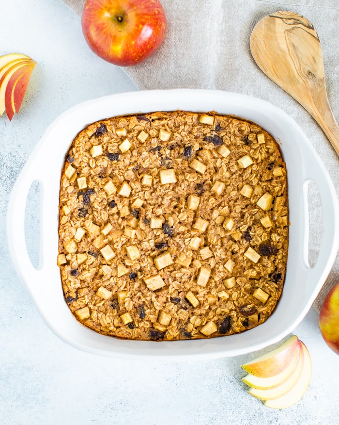 Dish of apple cinnamon baked oatmeal filled with raisins and chunks of apples. Apples and a wooden spoon are around the dish.