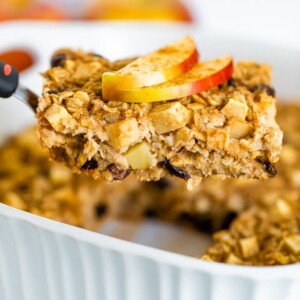 Spatula lifting a slice of apple cinnamon baked oatmeal from a baking dish. Oatmeal topped with apple slices and cinnamon.