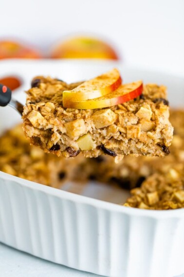 Spatula lifting a slice of apple cinnamon baked oatmeal from a baking dish. Oatmeal topped with apple slices and cinnamon.