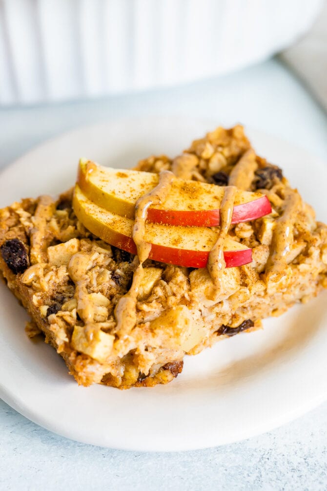 Slice of apple cinnamon baked oatmeal on a plate topped with apple slices and a nut butter drizzle.