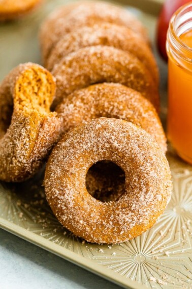 Apple cider donuts on a tray. One had a bit taken out of it, and a mason jar of cider is beside the donuts.