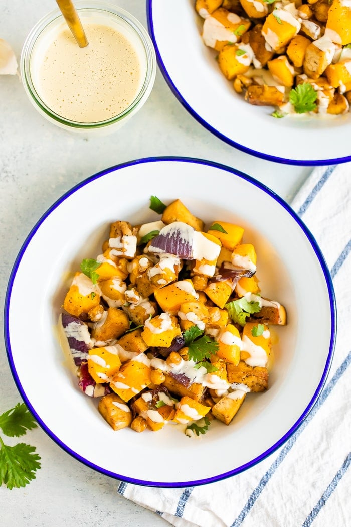 Two bowls of roasted butternut squash, tofu, chickpeas, and onion topped with cilantro and tahini sauce. A gold fork and jar of tahini sauce are on the side of the bowls.