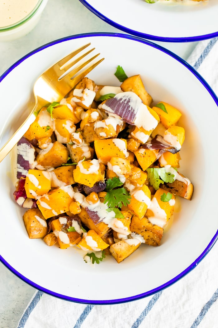 Bowl of roasted butternut squash, tofu, chickpeas, and onion topped with cilantro and tahini sauce. A gold fork is on the side of the bowl.