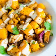 Bowl of roasted butternut squash, tofu, chickpeas, and onion topped with cilantro and tahini sauce. A gold fork is on the side of the bowl.