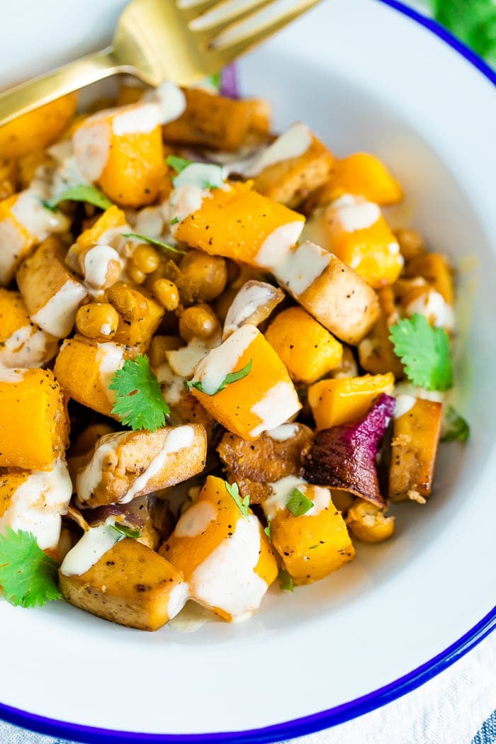 Bowl of roasted butternut squash, tofu, chickpeas, and onion topped with cilantro and tahini sauce. A gold fork is on the side of the bowl.