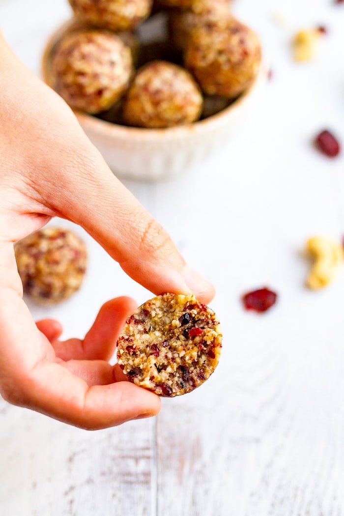 Hand holding a cranberry energy bite with a bite taken out of it. A bowl full of cranberry energy bites is in the background.