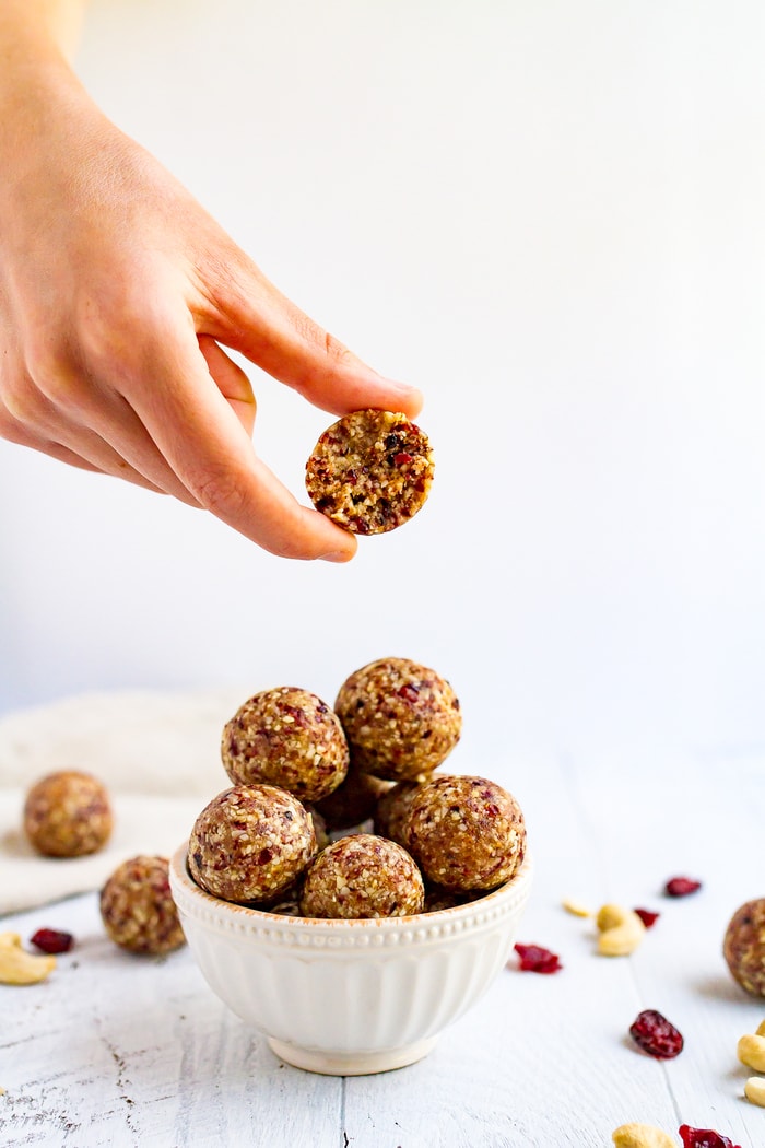 Hand holding a cranberry energy bite with a bite taken out of it. Hand is holding an energy bite above a bowl full of cranberry energy bites.