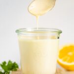 Glass jar with tahini dressing and a gold spoon drizzling some sauce off the spoon. Garlic, parsley and a lemon are around the jar.