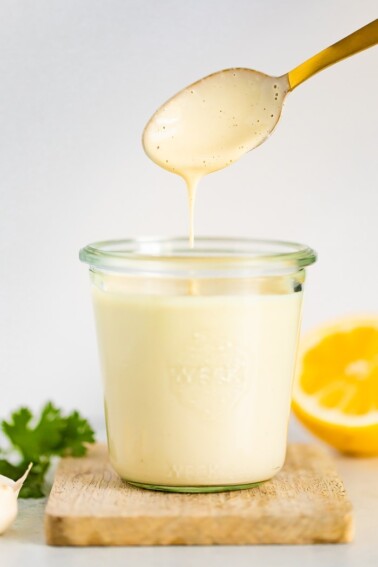 Glass jar with tahini dressing and a gold spoon drizzling some sauce off the spoon. Garlic, parsley and a lemon are around the jar.