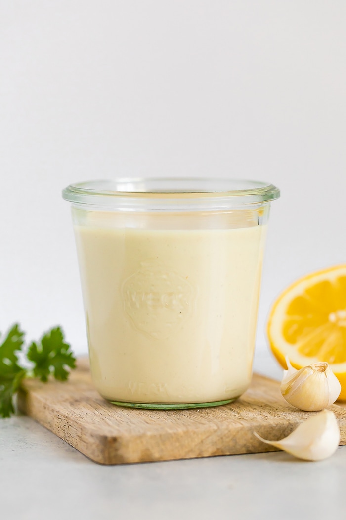 Glass jar with tahini sauce. Fresh parsley, half a lemon and garlic cloves are around the jar.