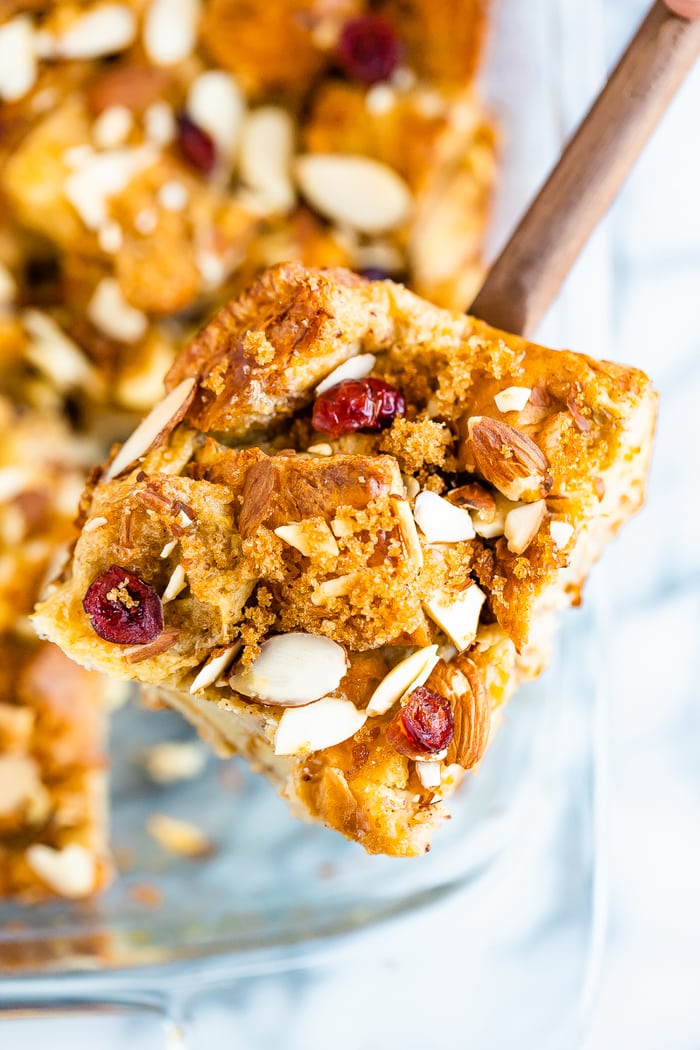 A slice of french toast bake on a wooden spoon with a casserole dish of the french toast back in the background. Bake is topped with cranberries and almond slices.