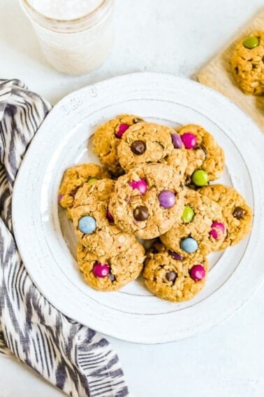 Plate of gluten-free peanut butter monster cookies with color candies in them. A dish towel and glass of milk are on the side.