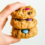 Hand holding a stack of three gluten-free peanut butter monster cookies made with oats and colored candies.