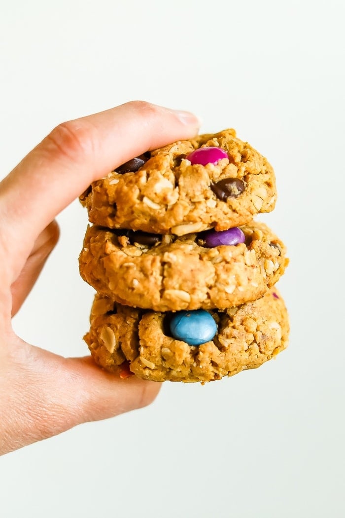 Hand holding a stack of three gluten-free peanut butter monster cookies made with oats and colored candies.