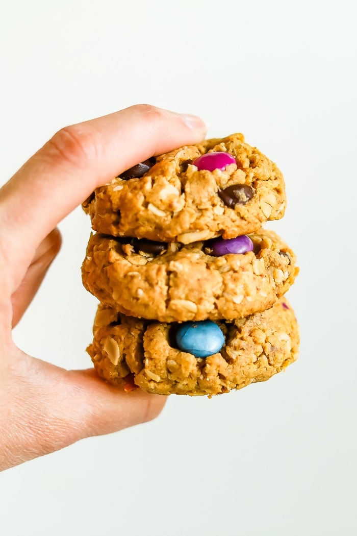 Hand holding a stack of three gluten-free peanut butter monster cookies made with oats and colored candies.