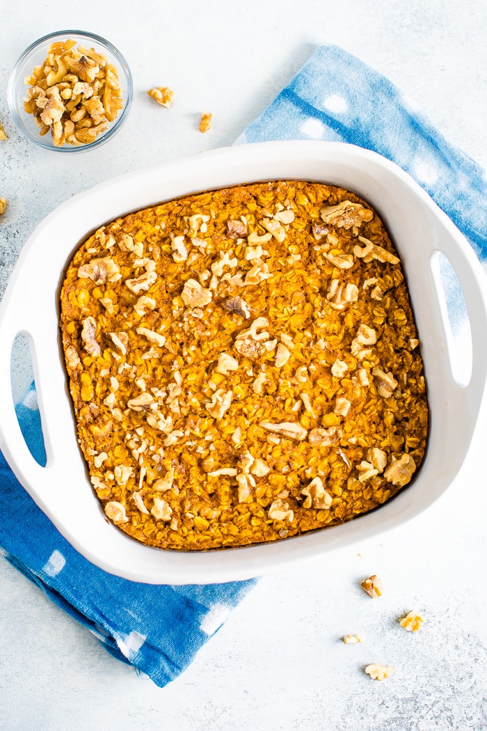 Square baking dish with pumpkin baked oatmeal topped with chopped walnuts. The dish is resting on a dish cloth and a bowl of walnuts is to the side.