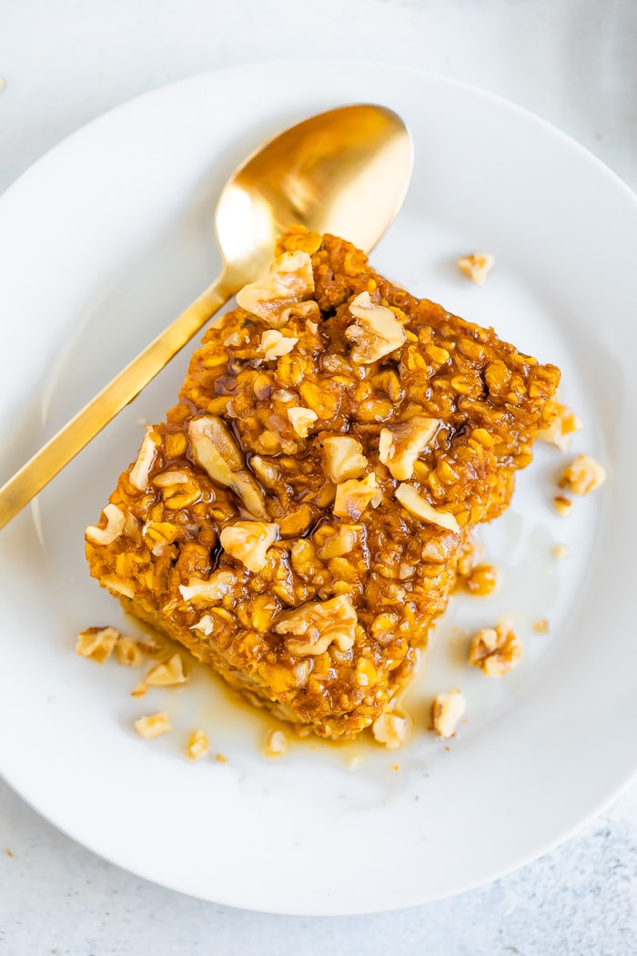 Slice of pumpkin baked oatmeal topped with walnuts and maple syrup. Oatmeal is on a plate with a gold spoon.