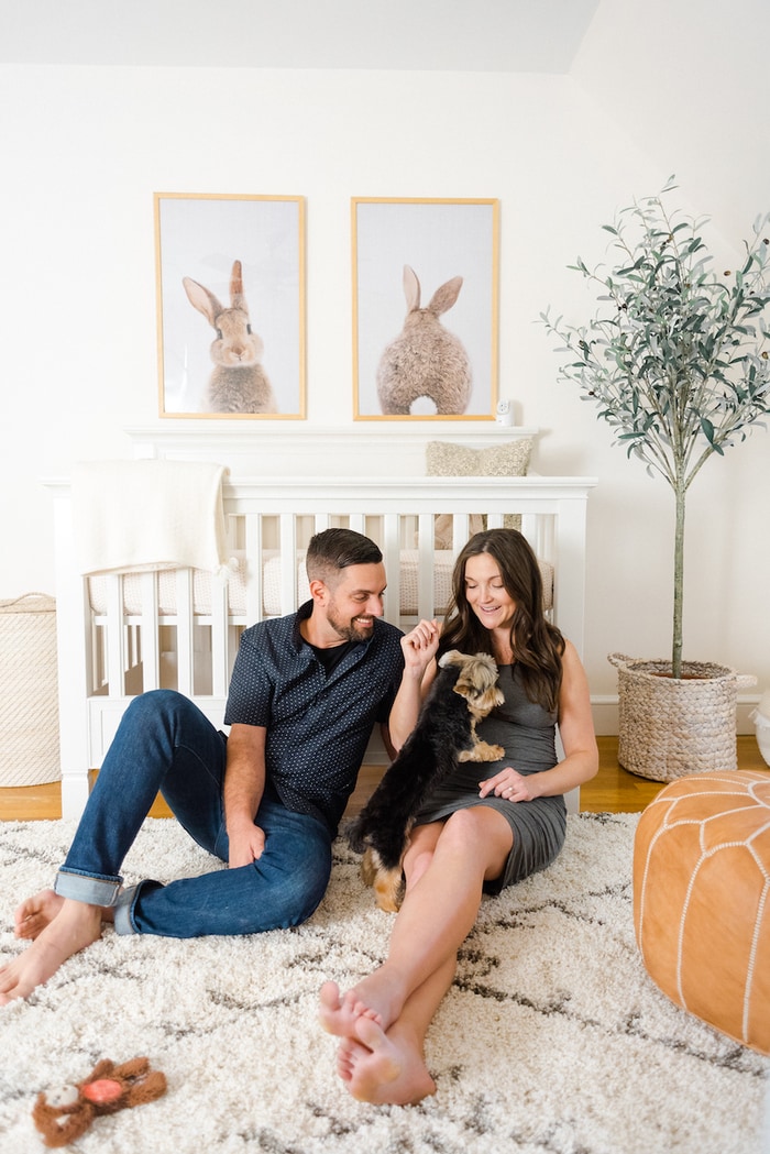 Expecting mom and dad sitting on the nursery floor with dog. 