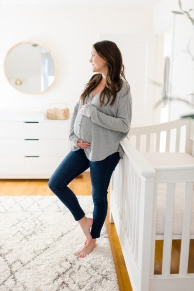 Expecting mom, leaning against crib and holding belly in the nursery.