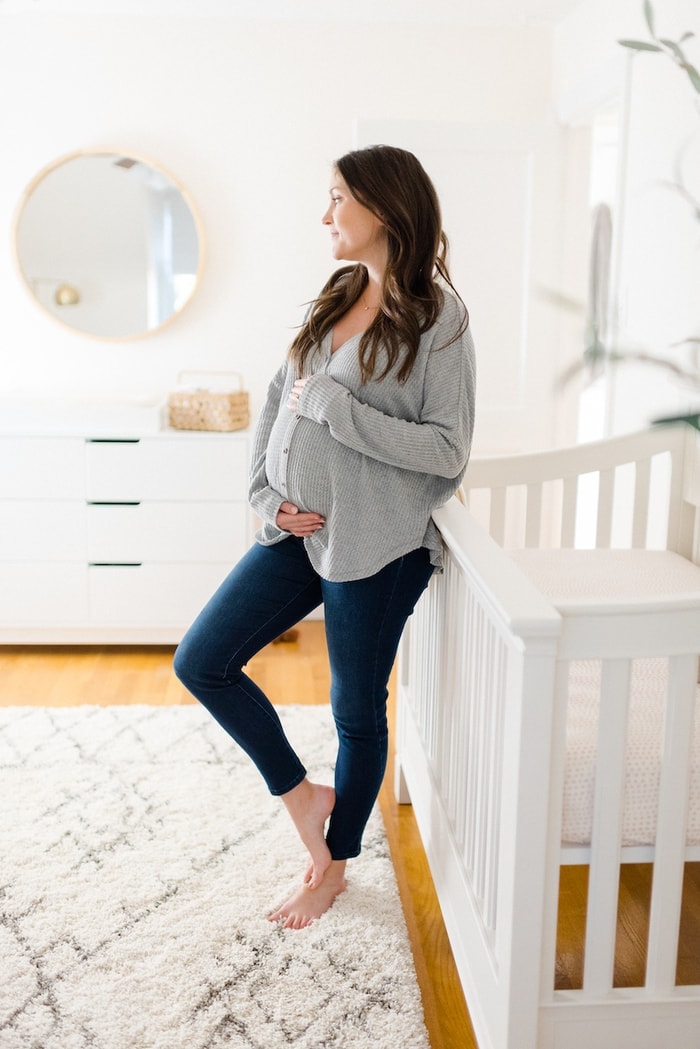 Expecting mom, leaning against crib and holding belly in the nursery. 