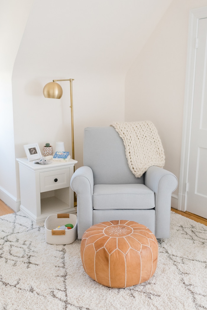 Baby girl nursery with a blue gray glider, gold flour lamp and leather pouf. 