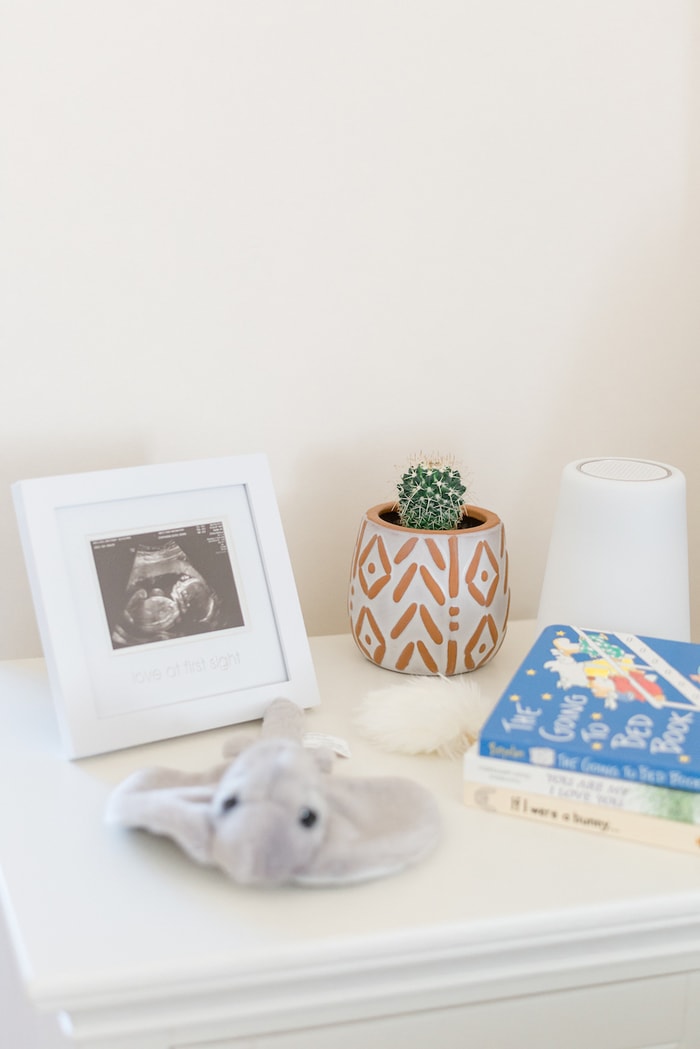 Nightstand in a nursery with sonogram photo, mini cactus, noise machine, stuffed stingray and books. 