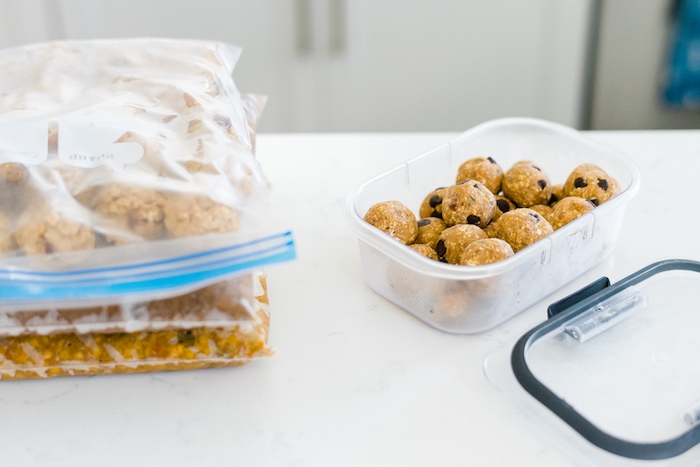 A plastic storage container with energy bites and three bags of frozen meal prep meals on a kitchen counter. 