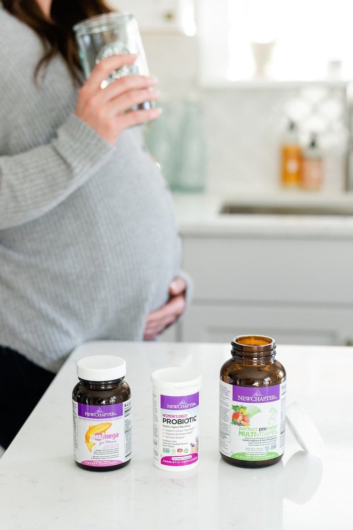 Pregnant woman holding her stomach and a glass of water. Three supplements are on the counter in front of her.