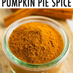 Small glass jar with pumpkin pie spice on a wood board and next to two sticks of cinnamon.