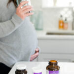 Pregnant woman holding her stomach and a glass of water. Three supplements are on the counter in front of her.