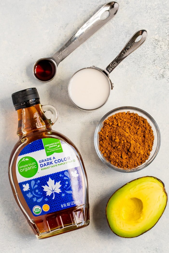 Ingredients to make avocado chocolate pudding: a teaspoon of vanilla, a measuring cup with almond milk, a bowl of cocoa powder, half an avocado, and a container of maple syrup.