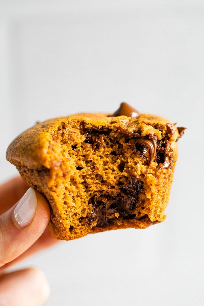 A woman's hand is holding up a flourless pumpkin muffin that has a bite taken out of it.