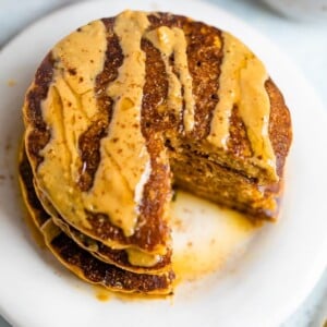 Stack of pumpkin pancakes, drizzled with peanut butter and maple syrup, with a bite taken out of the stack.