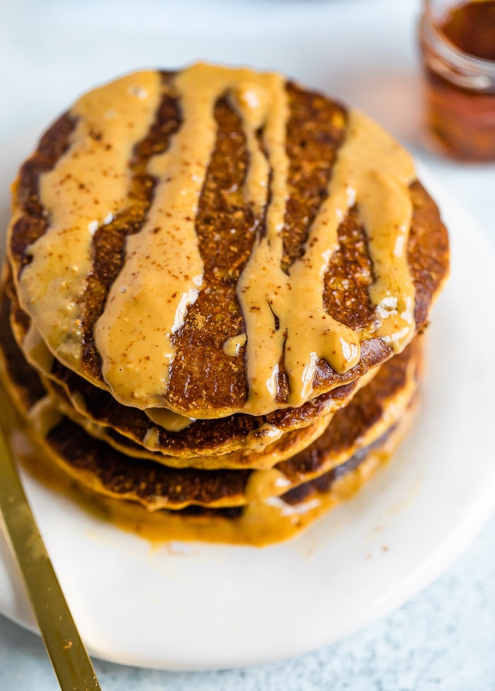 Stack of healthy pumpkin pancakes drizzled with peanut butter. A jar of maple syrup is beside the pancakes.