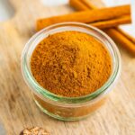 Small glass jar with pumpkin pie spice on a wood board and next to two sticks of cinnamon.