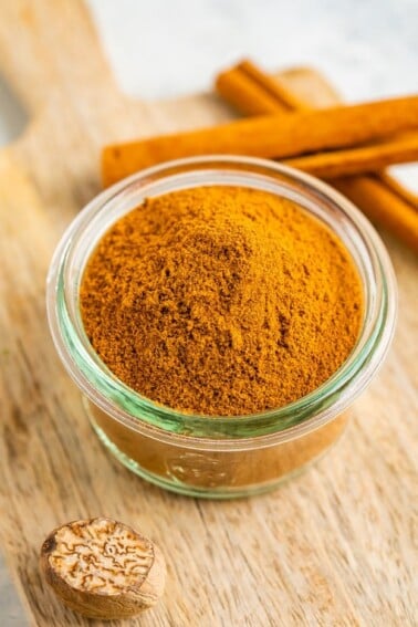 Small glass jar with pumpkin pie spice on a wood board and next to two sticks of cinnamon.