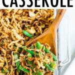 Casserole dish with healthy green bean casserole topped with crispy onions. A wood spoon is scooping some of the casserole.